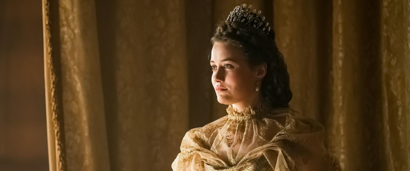 A young woman dressed in a gold gown and crown stands in front of gold damask curtains.