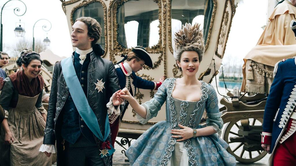 Louis XIV and Marie Antoinette hold hands in front of a guilded horse-drawn carriage.