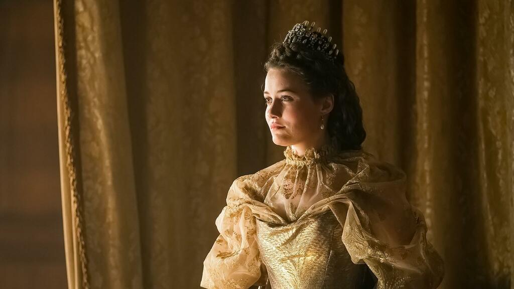 A young woman wears a golden gown and crown, in front of golden damask curtains.